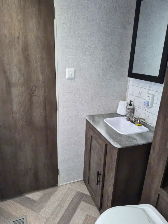 Clean and modern RV rental bathroom featuring a wood-look door, vanity with sink, mirror, and textured gray walls.. Forest River Wildwood 2021
