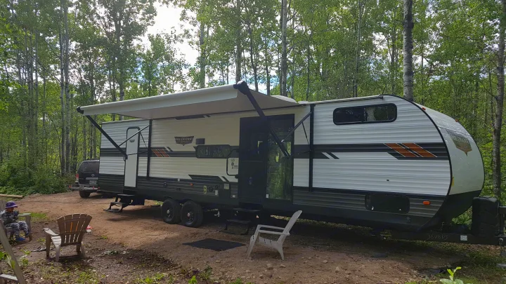 An RV rental, a white and grey travel trailer with an extended awning, is parked in a forest campsite where a child relaxes.. Forest River Wildwood 2021