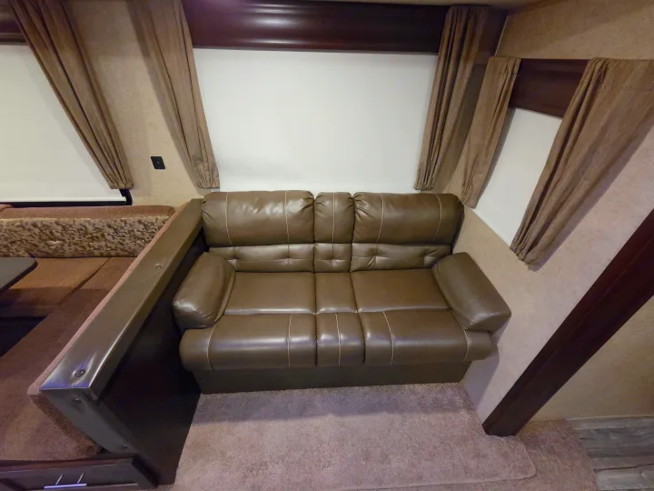 Spacious RV rental living area featuring a brown leatherette sofa with white stitching, brown curtains, and a dinette booth.. Forest River Cherokee 2017
