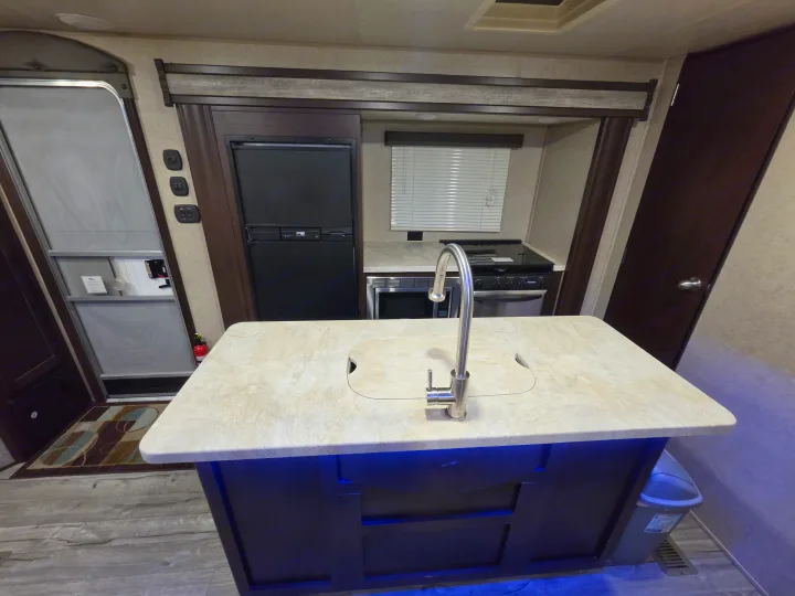 RV rental kitchen with a central island sink, under-cabinet blue lights, a refrigerator, stove, and microwave.. Forest River Cherokee 2017