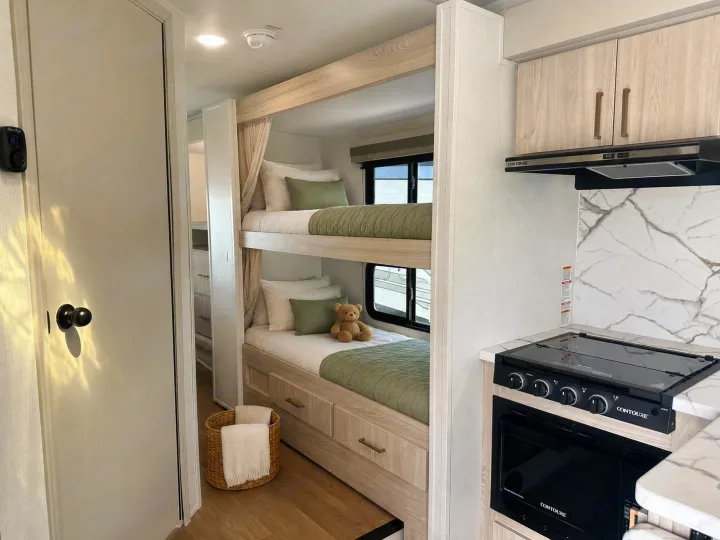 Modern RV rental bunk beds with sage green bedding and a compact kitchen area featuring a stove and oven. Ideal family space.. East to West Entrada 2026