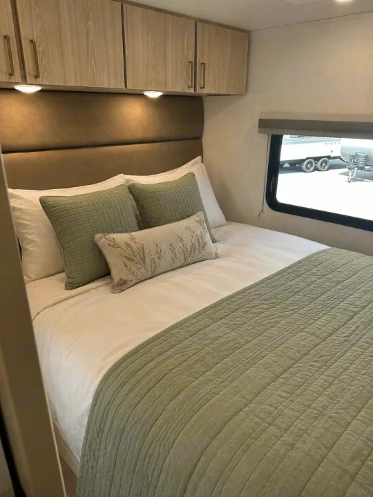 Cozy RV rental bedroom featuring sage green bedding, padded headboard, overhead storage, and a window view.. East to West Entrada 2026
