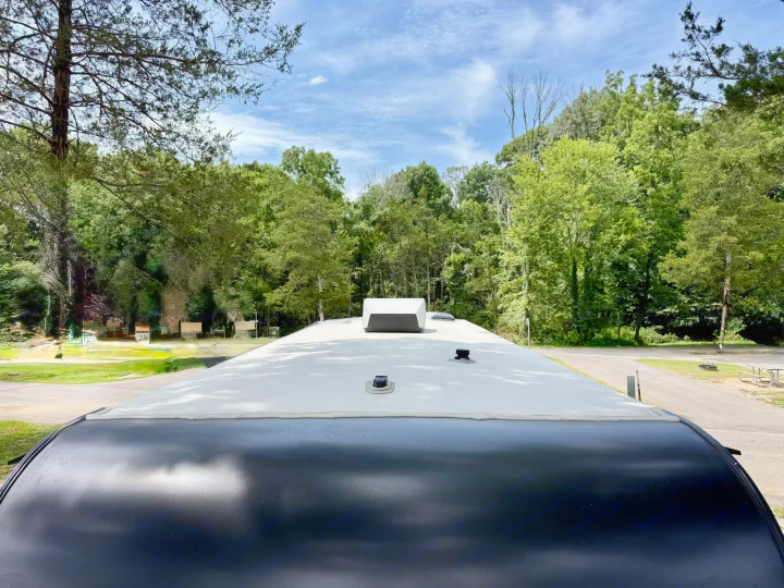 View from an RV rental roof at a wooded campground, showing the black front cap, white roof, sky, and surrounding trees.. Grand Design Transcend Xplor 2025