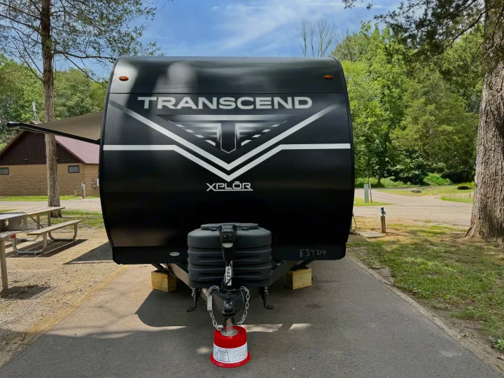 Black Transcend XPLOR travel trailer rv rental at a shaded campsite. Front view shows propane tanks and jack block.. Grand Design Transcend Xplor 2025