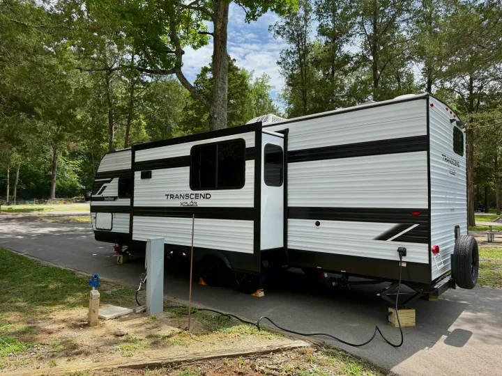 An RV rental, a Transcend Xplor travel trailer, at a shaded campsite with utility hookups connected.. Grand Design Transcend Xplor 2025