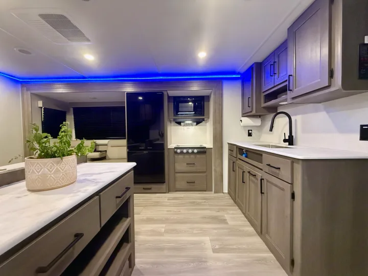 Modern RV rental kitchen interior with gray cabinets, black appliances, light counters, blue LED lighting, and a living area.. Grand Design Transcend Xplor 2025