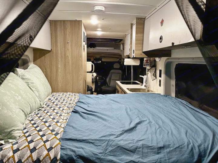Comfortable rv rental interior featuring a made bed with patterned bedding, a compact kitchen with sink, and overhead cabinets.. Winnebago Solis 59PX 2023