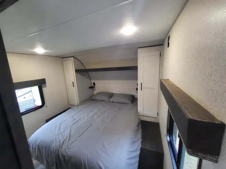 This RV rental offers a cozy bedroom with a neatly made gray bed, flanking cabinets, an overhead shelf, and windows.. KZ Sportsmen SE 332bkhse 2022