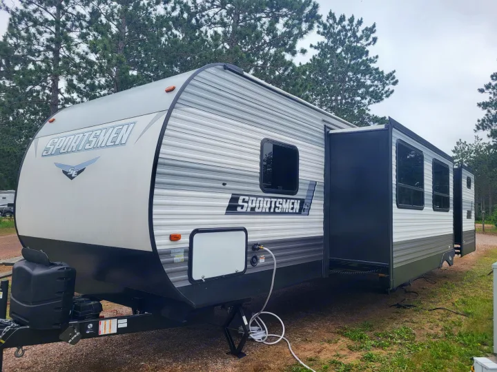 A grey and white Sportsmen travel trailer rv rental, with its slide-out extended, parked at a rustic campsite under pine trees.. KZ Sportsmen SE 332bkhse 2022