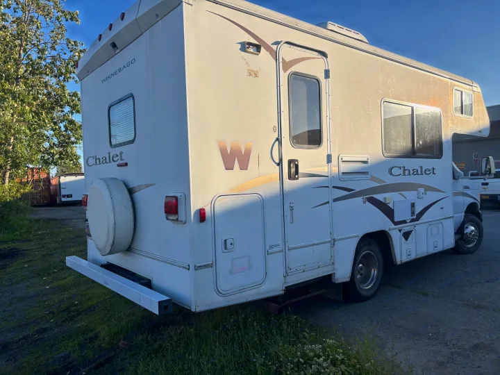 A weathered white Winnebago Chalet RV, ideal for rv rental, is parked outdoors, showing its rear, side, and spare tire.. Winnebago Chalet 2007