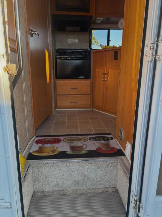 Step inside this RV rental's kitchen, featuring a stove, microwave, wood cabinets, and a welcoming coffee mat at the entry.. Winnebago Chalet 2007