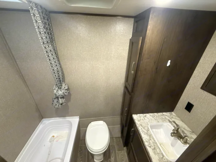 Compact RV rental bathroom featuring a shower with a patterned curtain, toilet, and sink vanity with dark wood cabinets.. Jayco Jay Flight SLX 2021