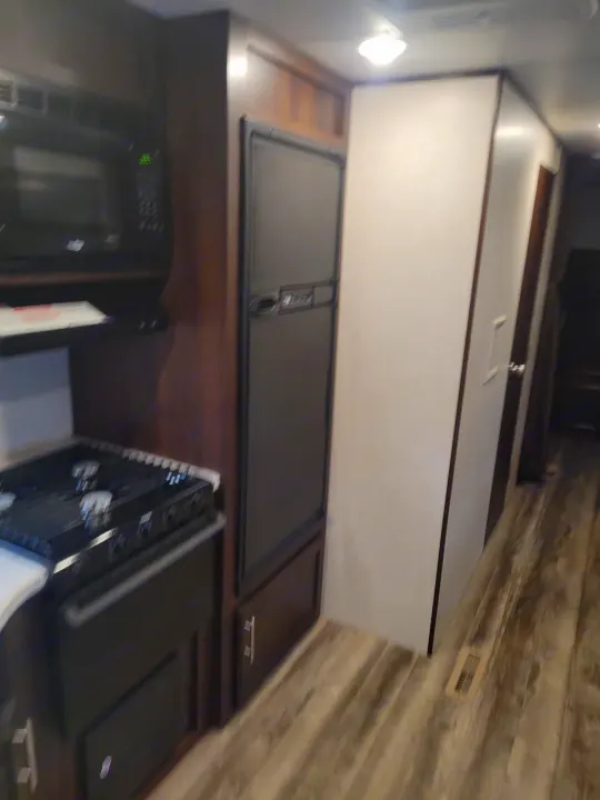 RV rental interior: Kitchen area with black microwave, stove, and refrigerator next to dark wood cabinets, light flooring.. Jayco Jay Flight SLX 2019