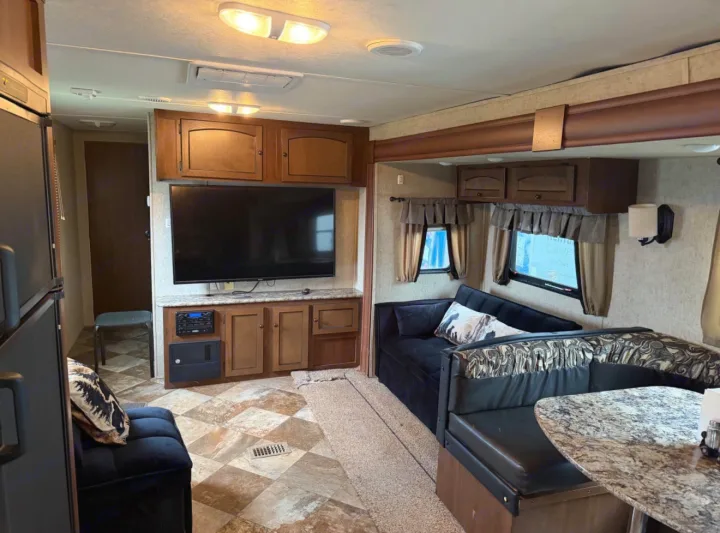 RV rental living space with a large TV, wood cabinets, black sofa, and patterned dinette. Cozy travel trailer interior.. Layton Layton Trailer 2014
