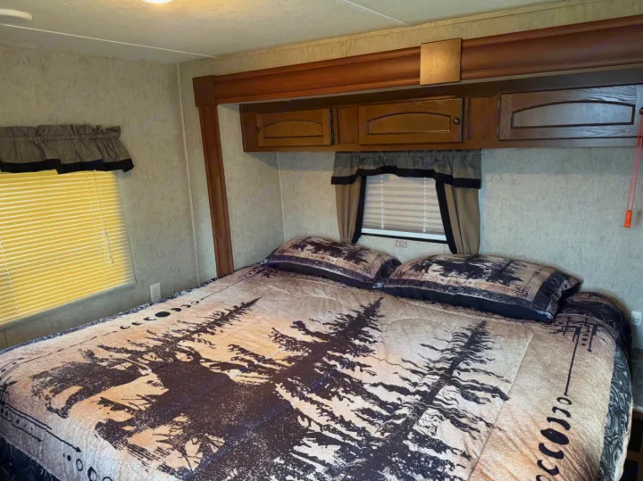 RV rental bedroom featuring a bed with forest-patterned bedding, overhead cabinets, and windows with blinds and valances.. Layton Layton Trailer 2014