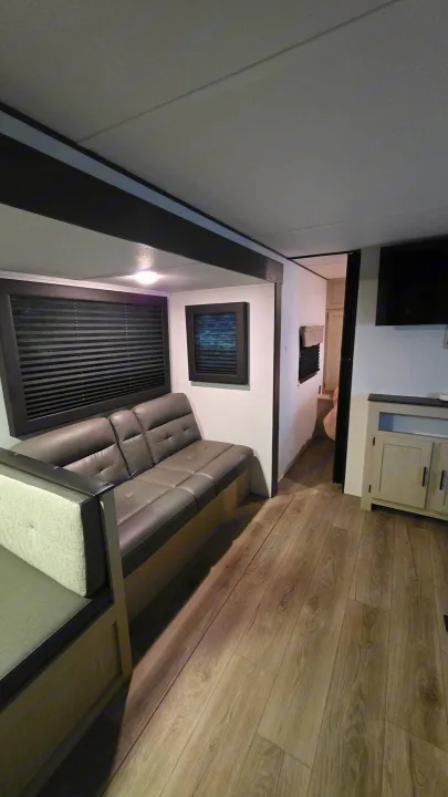Modern rv rental living area features a gray sofa, built-in dining bench, dark blinds, and wood-look flooring.. Forest River Shasta I-5 2025