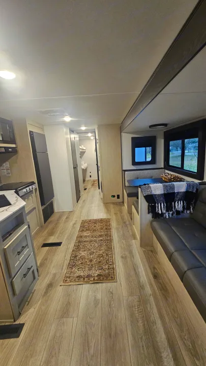 Modern rv rental interior: kitchen, dining nook with plaid blanket, long couch, light wood floor, and hallway to bunk beds.. Forest River Shasta I-5 2025