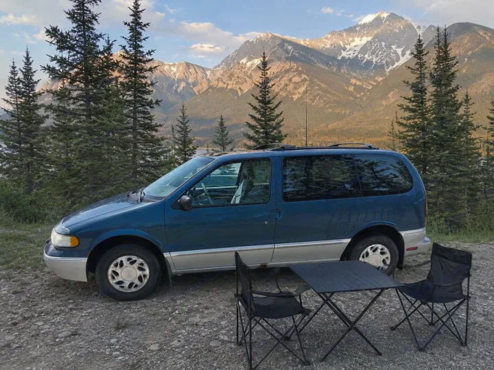 Blue minivan, camping table & chairs by majestic mountains. An ideal RV rental alternative for scenic trips.. Mercury Villager 1997