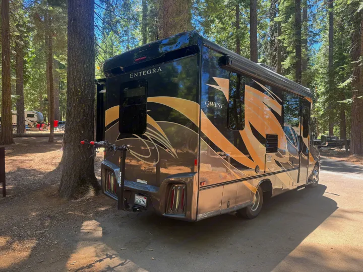 Black and gold Entegra Qwest rv rental with a bike rack, parked in a sunlit forest campground among tall trees.. Entegra Coach Qwest 2018