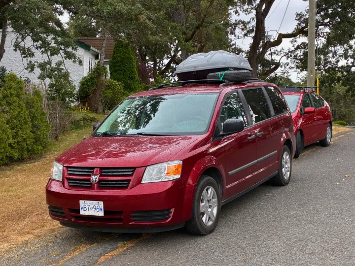 Red Dodge Grand Caravan, an RV rental alternative, with rooftop cargo & tire, parked on a damp road in BC.. Dodge Grand Caravan 2010
