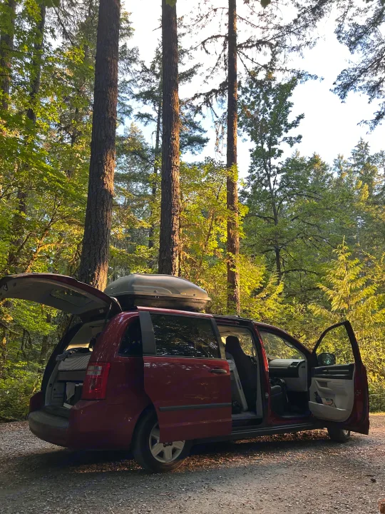 Red minivan, set up like an RV rental with a roof box & bed, ready for a forest camping adventure. Doors open in dappled light.. Dodge Grand Caravan 2010