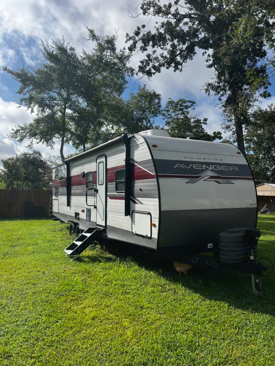 An RV rental, a white and maroon travel trailer, parked on a lush green lawn under a blue, cloudy sky. Surrounded by trees, with steps ready for boarding.. Avenger Avenger Trailer 2025