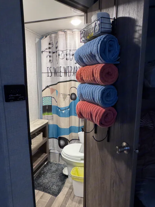 Compact RV rental bathroom with a themed shower curtain, toilet, wooden shelves, and rolled blue & coral towels on a door-mounted rack.. Crossroads RV Zinger Lite 2021