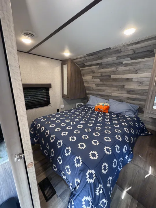 This inviting rv rental bedroom features a blue patterned comforter, wood accent wall, and ceiling lights, offering a comfortable camper interior.. Crossroads RV Zinger Lite 2021