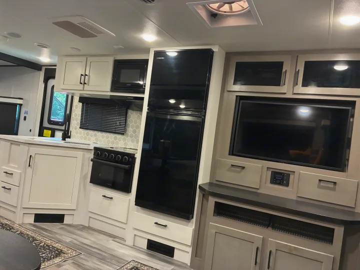 Modern rv rental interior: bright kitchen with black appliances, light cabinets, and entertainment area with TV.. Jayco Jay Feather 2024