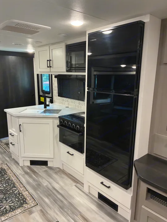 Modern RV rental kitchen: white cabinets, light counters, black Everchill fridge, Furrion microwave, and wood-look floor.. Jayco Jay Feather 2024