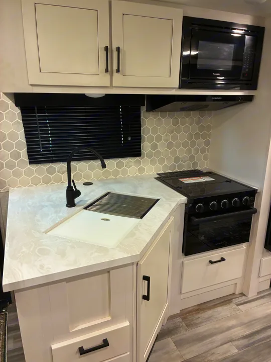 Modern RV rental kitchen with white cabinets, light marbled counters, black appliances, and a gray hexagonal backsplash.. Jayco Jay Feather 2024