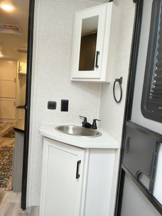This rv rental features a compact, modern bathroom with a corner sink, white cabinets, and a mirrored medicine cabinet.. Jayco Jay Feather 2024