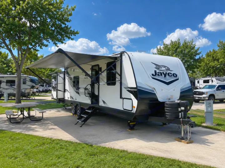 A Jayco RV rental travel trailer with an extended awning sits on a sunny campground site, complete with a picnic table.. Jayco Jay Feather 2024