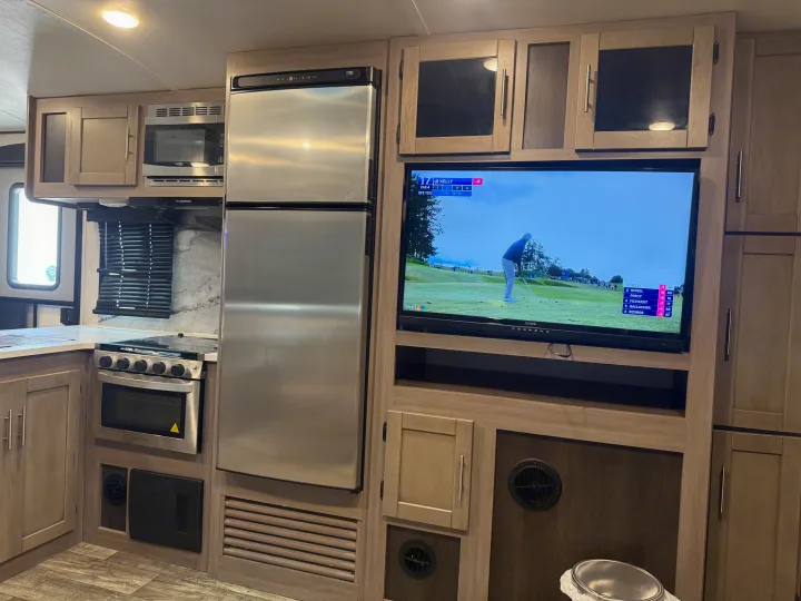 Modern RV rental interior featuring a kitchen with stainless steel fridge, microwave, oven, and a TV showing golf.. Crossroads RV Sunset Trail Super Lite 2022