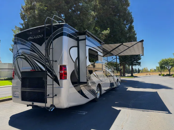Rear view of a modern silver and black Thor Palazzo motorhome RV rental with extended awning, parked in a sunny lot.. Thor Palazzo 2017