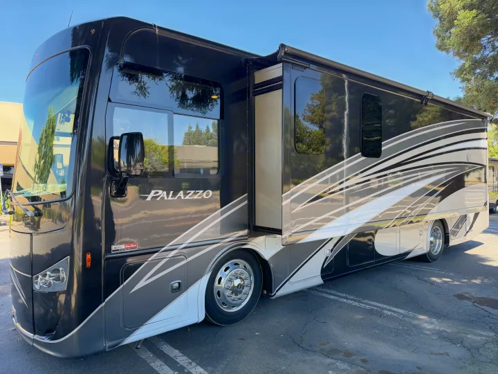 A large, black and white Palazzo RV rental is parked on asphalt with its side extended, reflecting trees.. Thor Palazzo 2017