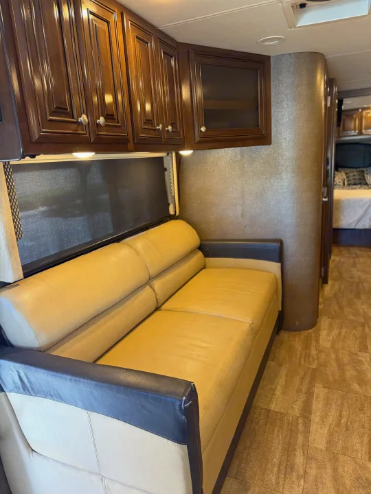 RV rental interior features a cozy beige sofa, rich wood cabinets, and patterned tile floor, with a bedroom visible in the background.. Thor Palazzo 2017