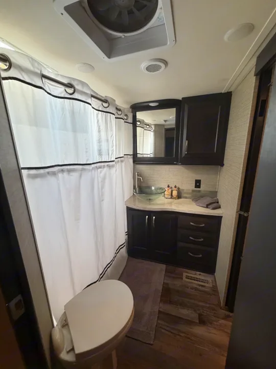 Compact RV rental bathroom showing a toilet, shower with curtain, dark vanity with glass vessel sink, and overhead fan.. Denali Denali Fifth Wheel 2017