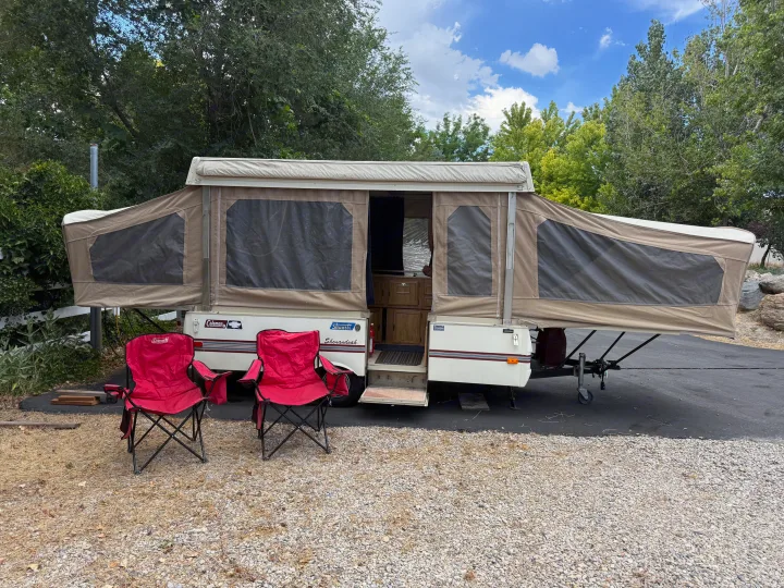 A fully set-up pop-up rv rental with two red chairs, ready for camping in a wooded outdoor setting.. Coleman Grand Tour Shenandoah 1987