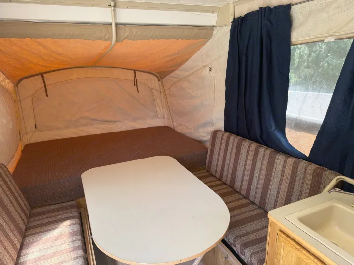 Cozy pop-up RV rental interior with dinette, bed, sink, and blue curtains, offering a compact living space.. Coleman Grand Tour Shenandoah 1987