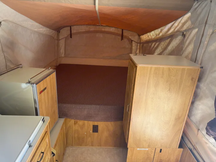 Cozy pop-up RV rental interior, featuring a bed with brown mattress, wooden cabinets, and a compact fridge.. Coleman Grand Tour Shenandoah 1987
