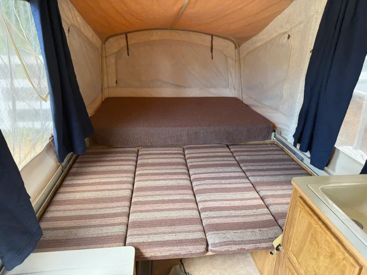 Inside an rv rental pop-up camper, showing a rear bed, a front convertible seating/bed area, blue curtains, and a sink.. Coleman Grand Tour Shenandoah 1987