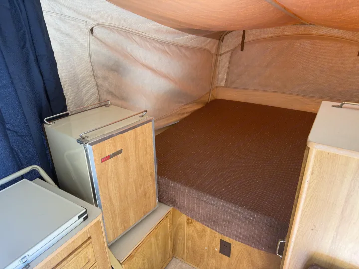 RV rental pop-up camper interior, showing a brown bed, vintage fridge, wood cabinets, and light fabric walls with mesh windows.. Coleman Grand Tour Shenandoah 1987