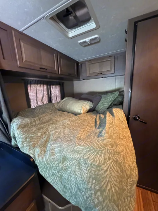 Compact RV rental bedroom featuring a comfy bed with green botanical bedding, dark wood cabinets, and overhead vent.. Thor Majestic 2018