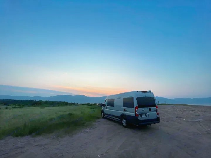 RV rental camper van parked on a sandy track at sunset, with mountains and a grassy field in the background. Ideal for outdoor travel.. RAM Dodge Promaster 2015