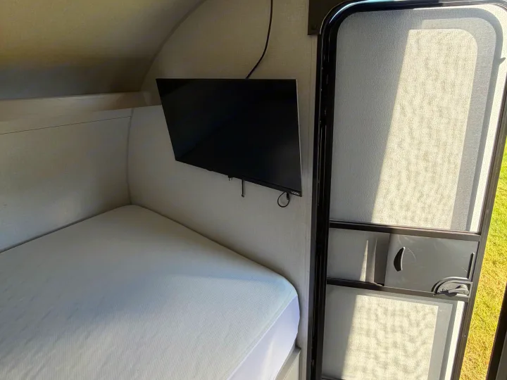 Cozy rv rental interior with a neatly made bed, wall-mounted TV, and an open screen door to the outside. An "EXIT" sign is visible.. Coachmen Clipper 17BH 2024