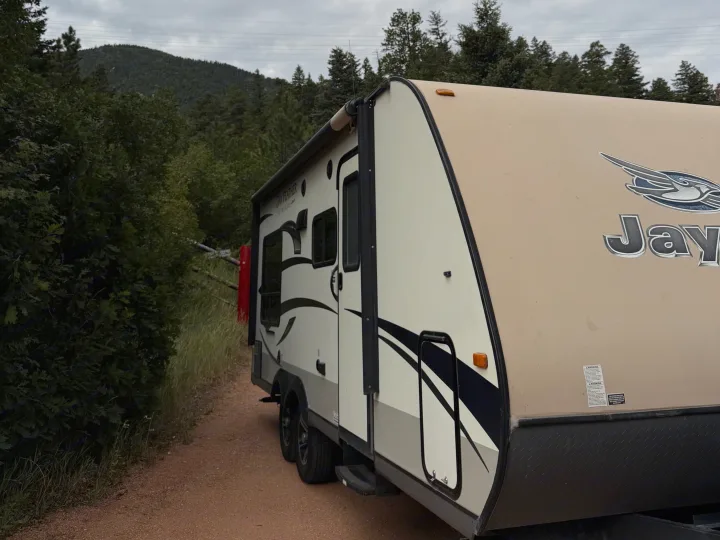 Jay Feather travel trailer rv rental parked on a dirt path, with green trees and mountains in the background under a cloudy sky.. Jayco Jay Feather Ultra Lite 2015