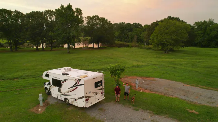 RV rental at a scenic campsite. Two men & their dog stand by their Class C RV next to a lake and green fields.. Thor Four Winds 2020