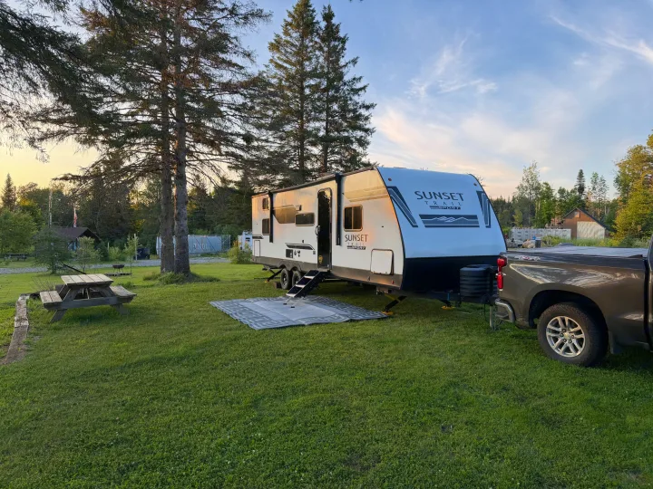 An rv rental: A Sunset Trail Sport travel trailer & truck parked at a green campground with picnic table, trees, at sunset.. Crossroads RV Sunset Trail Sport 2025