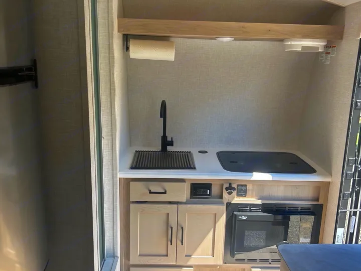 Modern RV rental kitchen with a black sink, cooktop, microwave, and light wood cabinetry.. Forest River Rockwood Geo Pro 2023